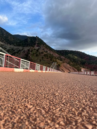 The image depicts a scene from Tabaroucht, Morocco, focusing on a textured wall or ground surface in the foreground, which appears to be made of small pebbles or rough material. Adjacent to the wall is a red and white guardrail extending into the distance. Beyond the guardrail, there is a mountainous landscape with some greenery and rocky terrain. The sky is visible in the background with a mix of blue sky and dark storm clouds. The image is rotated counter-clockwise.