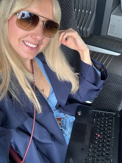 A young blonde woman is taking a selfie on a train. She is wearing sunglasses, a blue shirt, and a navy blue trench coat. A laptop is open on her lap. The background shows train seats. There is no indication of the city of Garupe, Latvia, in the image.