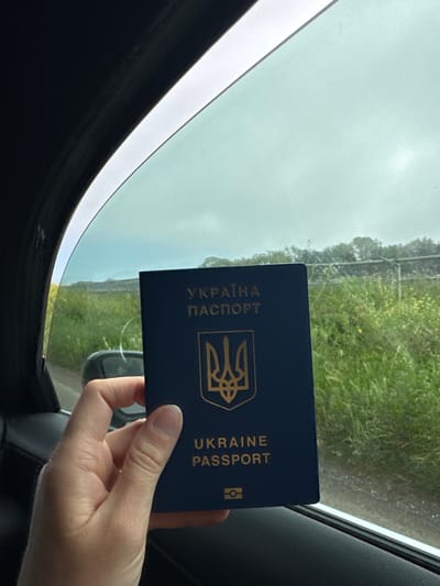 The image shows a hand holding a blue Ukrainian passport inside a car. The passport's cover displays the Ukrainian coat of arms and the words "УКРАЇНА ПАСПОРТ" and "UKRAINE PASSPORT" in gold lettering. The view through the car window reveals a blurred landscape with greenery and overcast sky. The photograph was possibly taken in the city of Cisterna di Latina, Italy.