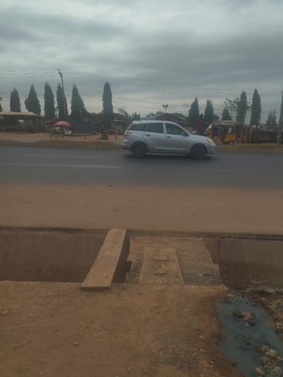 The image shows a silver minivan driving on a paved road. The road has a ditch on the right side. The ditch is partially filled with dirt and has a wooden plank bridge over it. The minivan is traveling in the direction of a small town in the distance. There are several trees lining the road. The sky is overcast. The scene is taken in Junction, Nigeria, at coordinates (latitude: 8.958, longitude: 7.551). There is a single person visible in the image, standing next to a yellow tricycle parked on the side of the road. The scene is a typical street in a rural area of Nigeria.