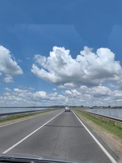 The image shows a gray asphalt road stretching into the distance under a blue sky with scattered white clouds. On the right side of the road, there is a metal guardrail and a grassy embankment. To the left of the road, there is a large body of water, likely a lake or river, bordered by a distant tree line. A silver car is driving away from the camera in the left lane. The scene appears to be in a rural or suburban area, with some buildings visible on the far shore of the water. The overall impression is one of a pleasant, sunny day for travel.