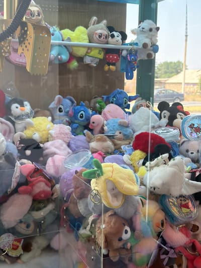 This image depicts a claw machine filled with an assortment of stuffed animals. The machine is placed outdoors, with a background showing a street, buildings, and trees, suggesting an urban or suburban setting. The context provided indicates the location as Verin Ptghni, Armenia. The stuffed animals are varied in color, size, and character, with notable examples including Stitch from Lilo & Stitch, a white sheep-like creature, a penguin, and what appears to be a brown teddy bear. The claw mechanism itself is visible at the top left, a metallic gripper. The lighting suggests it is daytime. There are no people or visible interactions with the machine in this particular frame.