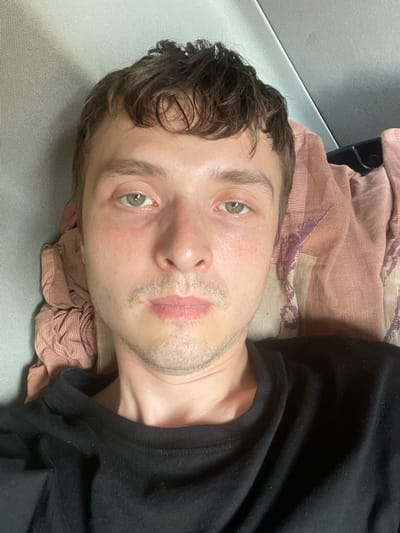 A person is lying down on their back on a textured surface, possibly a bed or couch. They are wearing a black t-shirt. A patterned textile, in shades of pink and beige, is visible beneath their head and shoulders. The person has light skin, light brown curly hair, and green eyes. There are freckles on their cheeks. Their lips appear slightly smudged, and there is a hint of stubble on their face. The lighting suggests it is daytime, but there are no clear indicators of the weather or specific location beyond the general context provided. The pose and expression could be interpreted as relaxed or perhaps a bit weary. No text is visible in the image.