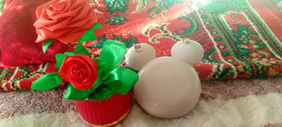 This image depicts a still life arrangement on a surface that appears to be a textured rug, with a smaller, fluffy textile beneath it. The primary focus is on decorative items. In the foreground, to the left, is a small red container with a golden rim, filled with a faux red rose and green ribbon. To the right, a large, smooth, light-colored object resembling a Mickey Mouse head with two smaller spherical "ears" is prominently featured. In the background, a larger, folded red textile is visible, partially overlapping a red, green, and yellow patterned rug. The lighting suggests an indoor setting with natural light coming from the upper right, casting shadows and creating bright highlights. There is no indication of the city of Marhamat, Uzbekistan, or any elements that would geographically place the scene there.