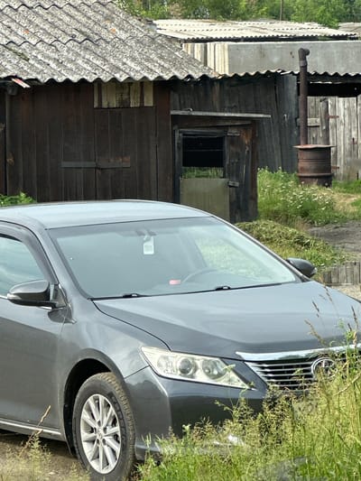 In the foreground of the image is the front passenger side of a dark gray sedan, likely a Toyota Camry, partially obscured by tall green grass and wildflowers. The car's headlights and grille are visible. Behind the car, a weathered wooden structure with a corrugated roof stands. This structure appears to be a shed or outbuilding, with a dark, vertical wooden door. To the right of this building, a metal barrel is situated, with a tall, rusty metal pipe extending upwards from it, resembling an old stove or incinerator. Beyond this, another wooden structure with a corrugated roof is visible. The background suggests a rural or semi-rural setting, possibly an industrial yard or a collection of utility buildings, consistent with a location in Yermakovskoye, Russia. The overall scene depicts an outdoor environment with signs of age and disuse.