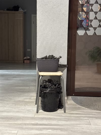 The image shows a chair with a black wastebasket hanging from it, positioned against a wall near a window. The chair appears to be a small, utilitarian model with a light-colored seat. The wastebasket is overflowing with black plastic bags. The background includes a cabinet and a partial view of a window with a patterned design. The flooring is light-colored wood or laminate. The specific location within the city of Zamankul, Russia is not discernible from the image. No people are visible.