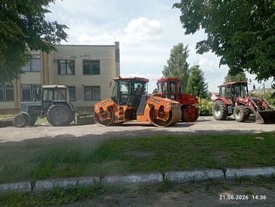 In the city of Хаценчыцкі сельскі Савет, Belarus, on a paved surface in front of a low-rise brick building with visible signs of wear, four construction vehicles are parked. The leftmost vehicle is a blue tractor with large rear wheels and a smaller front set. To its right is an orange road roller, a piece of heavy machinery with two large drums, one at the front and one at the rear. Next to the orange roller is another red road roller, similar in design. The furthest vehicle to the right is a red tractor equipped with a front-end loader. The scene is illuminated by daylight, with a partly cloudy sky and lush green trees providing a natural backdrop. The ground is a mix of asphalt and gravel, with a concrete curb running along the grassy foreground. A man is visible inside the cabin of the orange road roller. The timestamp "21.06.2026 14:36" is present in the bottom right corner of the image.