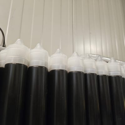 The image displays a row of identical black plastic bottles with clear, ridged caps, tightly packed together. The bottles are arranged in a horizontal line, with their caps facing upwards and slightly towards the viewer. The background consists of a vertically ribbed, off-white wall, possibly made of corrugated metal or plastic sheeting, which is illuminated from the left, casting subtle shadows between the ribs. The lighting creates a somewhat sterile, possibly industrial or storage environment. There are no people or animals visible in the image. The scene appears to be a close-up shot focusing on the bottles and the immediate background.