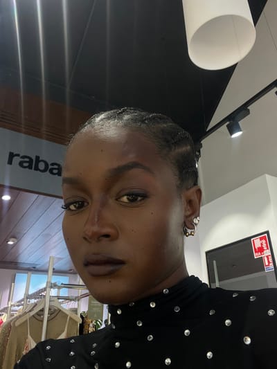 An individual, identified as a female with a dark complexion, is positioned centrally in a close-up, chest-up shot. Her dark hair is styled in close-set braids or cornrows. She has prominent eyebrows, eye makeup, and dark-toned lipstick. Small, scattered dark spots are visible on her forehead and cheeks. She wears multiple earrings on her visible left ear: a silver-toned hoop with two small gold-toned beads, and a smaller stud or hoop with clear stones. Her attire consists of a black turtleneck top adorned with numerous clear, round rhinestones or studs. She maintains direct eye contact with the camera.

The scene is an indoor commercial environment. In the upper left background, a light-colored sign displays the partial, capitalized, sans-serif text "raba" against a darker background. Below the sign, a clothing rack holds various garments, including one with sequined or embroidered detailing. A section of a wall featuring horizontal wooden slats is visible behind the clothing rack. The ceiling above is dark, equipped with multiple recessed spotlight fixtures and a large, white cylindrical pendant light fixture. To the right, a white wall segment is visible adjacent to a dark-framed opening or display. A red and white fire safety sign, indicating "RIA" (Robinet d'Incendie Armé - equipped fire hose reel) and an arrow, is mounted on the white wall.

The location is a retail establishment in Serris, France, consistent with a store found within a shopping center, such as the Val d'Europe.