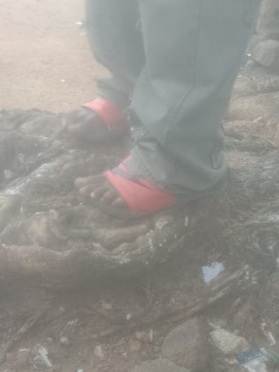A person's legs and feet are visible from the knees down, standing on rocky terrain. The person is wearing light-colored pants and red sandals with a dark accent. The sandals appear to have a Nike logo. The background is blurry, with a sandy area to the left and more rocks and debris to the right. The lighting suggests an outdoor environment. There are no other people or vehicles visible. The specific location within the city of Unknown, Unknown cannot be determined from the image.