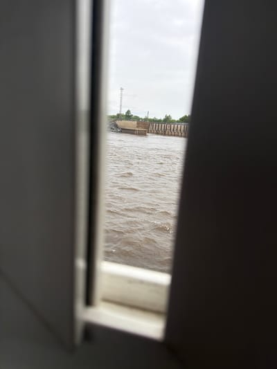 The image presents a view seen through a window or opening, framing a wide, muddy-colored river and its distant bank under an overcast sky. In the immediate foreground, dark, possibly metallic or plastic, horizontal bars form the top and bottom edges of the frame, obscuring much of the interior space from which the photo is taken. A blurred, lighter-colored window frame or interior structure is visible on the far right.

The visible outdoor scene is dominated by a body of water that appears turbulent with numerous ripples and small waves, reflecting the cloudy sky with muted highlights. The water's color is a murky brown or beige, characteristic of a sediment-rich river.

On the left side of the visible outdoor scene is the distant riverbank. A substantial embankment constructed from concrete or large stone blocks forms the shoreline, appearing as a series of rectangular segments. Above this structure, sparse green foliage, likely trees or bushes, can be seen, indicating some vegetation on the bank. Further along the bank, a tall, slender metal tower or mast stands against the bright, featureless sky. Dark riprap rocks are visible at the very base of the embankment, near the water's edge.

The sky is uniformly overcast and bright, suggesting daytime without direct sunlight. There are no people visible in the image, nor is any text discernable. The scene depicts a quiet, observational moment from an enclosed space, overlooking what appears to be a significant riverine environment, consistent with the location context of Nizhneleninskoe, Russia, and the Amur River.