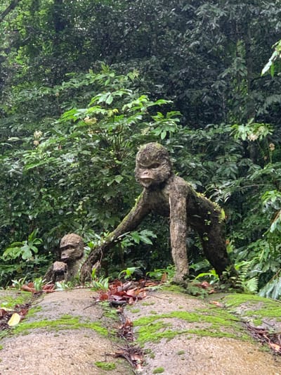 Two statues resembling apes, constructed from a material that mimics bark and moss, are positioned on a moss-covered rock formation in a lush, green forest. The larger ape statue is in the foreground, on the right, in a quadrupedal stance with its head turned to the left. Its body is covered in moss, giving it a natural, aged appearance. A smaller ape statue is visible to the left of the larger one, partially obscured by the foreground statue and appearing to be seated. The background consists of dense foliage and trees, suggesting a natural, tropical environment, possibly within Thôn 14, Vietnam, known for its abundant green spaces. The lighting is diffused, indicative of overcast conditions or a shaded forest canopy.