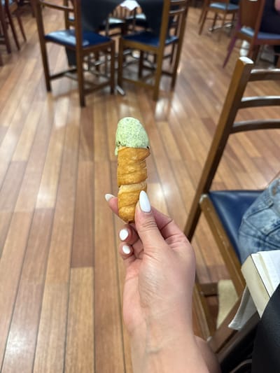 A person's hand with white-painted fingernails holds a cannoli with pale green filling. The cannoli appears to be a fried pastry shell filled with a creamy, textured substance, possibly pistachio ice cream or a similar dessert. The background is a warm-toned wooden floor with a visible grain pattern, suggesting an indoor setting. In the blurred background, there are several wooden chairs with dark blue seats, indicative of a cafe, restaurant, or similar establishment. The lighting suggests it is daytime. There is no visible indication of the location being in Torrejón de Ardoz, Spain.