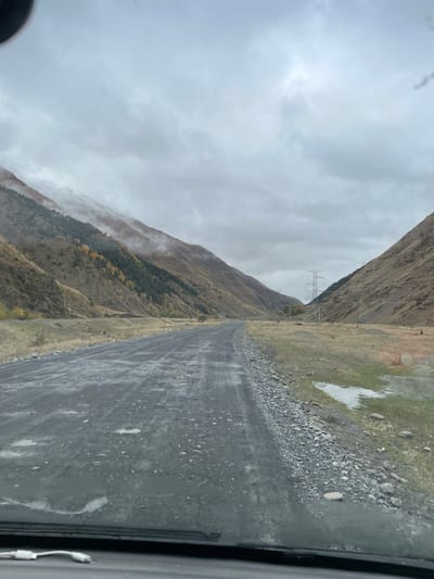 This image depicts a mountainous valley scene, likely near Karkucha, Georgia, under an overcast sky. The foreground reveals a view from inside a vehicle, with the lower edge of the windshield and a white cable (possibly a charging cable) visible. A wide, unpaved gravel road, showing wet patches and puddles, recedes into the distance, curving slightly to the left.
On the left side, a steep mountain slope rises, partially obscured by low-hanging clouds or mist near its upper reaches, where faint traces of snow are visible. The slope is covered with a mix of evergreen trees and deciduous trees displaying yellow foliage, indicating autumn or early winter. Further down, the vegetation is predominantly dry, brown grass and scrub. A faint, low wall-like structure is visible on the lower left slope.
On the right side, another mountain slope parallels the road, covered primarily in dry, brown, and ochre vegetation. The valley floor between the road and this slope is a flat expanse of dry grass with several visible puddles. In the distance, within the valley, a large metal lattice-type electrical transmission tower (pylon) is prominent, with power lines extending across the landscape. The sky is uniformly grey and heavily clouded, obscuring the upper portions of the mountains. No human or animal subjects are present.