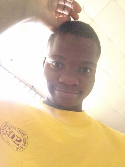 A brightly lit, low-angle selfie shows a young, dark-skinned individual, likely male, with short, dark hair. The person, positioned centrally in the frame, has a slight, friendly smile and appears relaxed. One hand is visible near the top of their head, seemingly either posing or adjusting hair.

The subject is wearing a vibrant yellow t-shirt. On the left side of the t-shirt, a circular emblem is clearly visible. This emblem contains Chinese characters and numbers. Prominently featured are the numbers "302". Surrounding text in Chinese translates to "National primary and secondary school students" and "Physical fitness exercises/calisthenics," indicating the t-shirt likely pertains to a student fitness program or event. The year "2013" is faintly discernable within the emblem, along with the letters "ASOS" or "ASOJ".

The background is an interior setting, characterized by a light-colored ceiling with visible tiles or panels. A very bright, possibly artificial, overhead light source illuminates the scene, casting a strong glow and creating an overexposed effect in parts of the image. The photo was taken indoors in Eleweran, Nigeria.