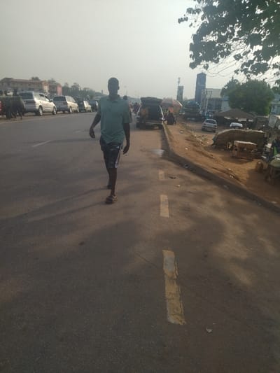 The image shows a man in a green shirt and black shorts walking on a paved road in Maraba, Nigeria, a city located at latitude 9.024, longitude 7.573. The man is walking toward the camera on the right side of the road, which has a yellow line painted down the middle. The man is in the center of the image, and his shadow is cast to the left of him. The image is taken from a street-level perspective. There are buildings and trees in the background, as well as a few parked cars, and a line of people walking on the opposite side of the road. There are some shops on the other side of the road, near the curb. The sky is hazy and the sun is low in the sky. The scene is a typical street scene in a Nigerian city.