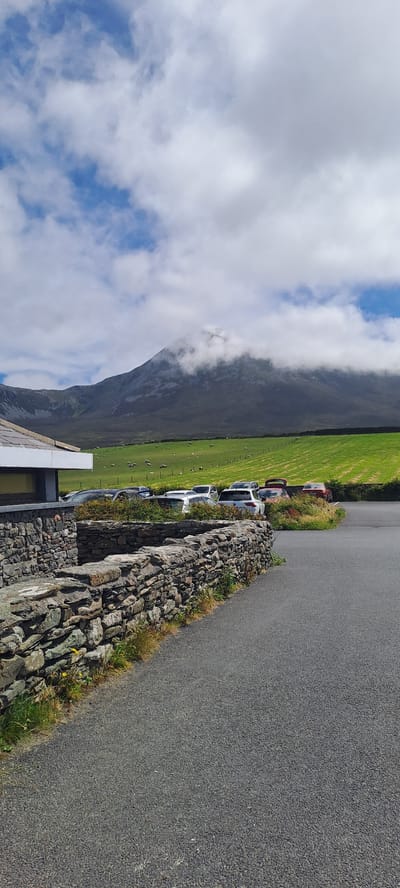 This image depicts a scenic view of a rural landscape, likely in Ireland. The scene is dominated by a dramatic mountain range partially obscured by clouds and fog, with a bright green, grassy field stretching out in front of it. A flock of sheep grazes peacefully in the field, adding to the pastoral atmosphere.
In the foreground, a paved road runs alongside a rough, stone-built wall. Several cars are parked along the edge of the field, suggesting a visitor or parking area. The cars appear to be a mix of sedans and SUVs, with some red and white vehicles visible. The time of day appears to be daytime, with bright sunlight casting shadows, and the weather seems to be mixed, with patches of blue sky peeking through fluffy white clouds.
The overall impression is one of natural beauty and tranquility, with a touch of human presence in the form of the parked cars and the road. The textures of the rough stone wall, the smooth asphalt of the road, and the soft green of the grass create a varied visual landscape. The lighting and cloud cover suggest a dynamic weather pattern, common in mountainous or coastal regions. No visible text is present in the image.