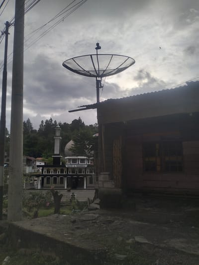The image captures a satellite dish positioned in front of a building with a corrugated metal roof, silhouetted against a cloudy sky. In the background, a mosque with a minaret and dome is visible, along with trees and other buildings. A utility pole stands to the left of the satellite dish. The scene appears to be an outdoor environment, possibly in a town or village in Blang Bebangka, Indonesia. The lighting suggests it may be daytime, but the overcast sky creates a somewhat somber atmosphere. No people are clearly visible or interacting in the frame.