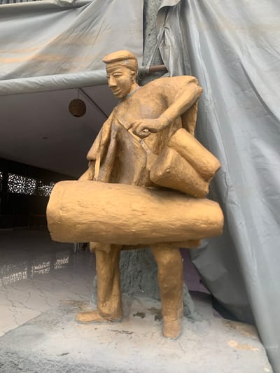 A gold-painted statue of a man wearing a cap and traditional attire is positioned in front of a grey tarp and a patterned wall. The statue depicts the man carrying bundles, one large one around his waist and two smaller ones on his shoulder. The background reveals a tiled floor and what appears to be an interior space with tables and chairs. The statue is located at the entrance to the Catholic Secretariat of the Catholic Bishops Conference of Nigeria in Bwari, Nigeria.