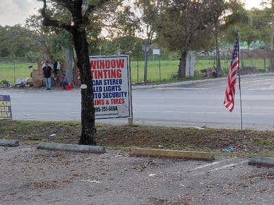 The image depicts an outdoor scene in Cutler Bay, United States of America. In the foreground, a dark, textured ground, likely asphalt or gravel, is covered with scattered brown leaves. Two concrete parking barriers are visible; one gray, and one yellow. A partial blue and yellow sign, reading "RESH" and "SH DAILY," is in the lower left corner.
A grassy median runs horizontally across the midground, separating the foreground from a two-lane asphalt road. A tree with dark, rough bark stands within this median, slightly left of center. To the right of the tree, a prominent white rectangular sign displays services in red and blue text: "WINDOW TINTING," "CAR STEREO," "LED LIGHTS," "AUTO SECURITY," and "RIMS & TIRES." A phone number "305-251-6668" and a website "autotronicsdrpot" are also visible, along with two QR codes. Further right in the median, an American flag is displayed vertically, hanging from a dark metal shepherd's hook.
Across the asphalt road, in the background, two individuals are visible near a cluttered area consisting of cardboard boxes, white plastic bags, and other debris. One person stands, wearing a dark shirt and blue jeans, facing right. Another person is seated on a red stool or bucket, wearing a blue cap and a striped shirt, facing left. A dark bicycle leans against the accumulated items. A wire fence runs behind this area, separating it from a greener, undeveloped lot. Various trees and shrubs populate the background, beneath an overcast or twilight sky.