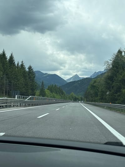 A view from inside a moving vehicle driving on a multi-lane highway. The highway has white lane markings and a metal guardrail. The sky is overcast with gray clouds. In the distance are mountains covered in green trees. The location is likely in a mountainous area near Pontebba / Ponteibe / Pontafel / Tablija, Italy, due to the mountain scenery and highway infrastructure.