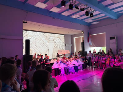 In Pron'kino, Russia, a group of children, likely students, are gathered in an indoor assembly hall, possibly for an event or performance. They are predominantly wearing blue and white scarves, suggesting a uniform or affiliation. Many of the children are holding lit candles and triangular signs with Russian text. The backdrop features a large screen displaying what appears to be vintage handwritten letters. The hall itself is illuminated with vibrant pink and purple stage lighting, and several spotlights are suspended from the ceiling. The floor has a patterned design. In the background, adults are also present, some observing the children and others taking photographs. A Russian flag is visible hanging on the wall. The overall scene suggests a solemn or commemorative occasion.