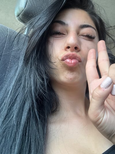 A person is posing for a selfie. The person is making a kissy face and holding up two fingers in a peace sign. The person has long dark hair and is wearing a denim jacket. The background is out of focus. It is impossible to determine if this image was taken in Strumyani, Bulgaria.