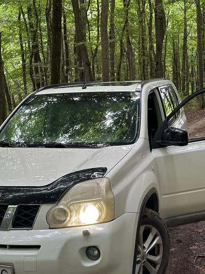 A white SUV is parked on a dirt road in a forest, presumably near Kolubaivtsi, Ukraine. The front of the vehicle is visible, with its headlights on. The driver's side door is open, and the interior is dark. The surrounding environment is a dense forest with tall trees and green foliage. The ground is covered in dirt and fallen leaves. The image appears to be taken from a low angle, looking up at the vehicle. There are no people visible in the image.