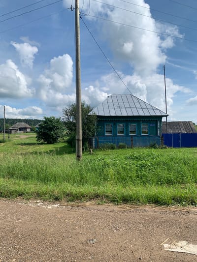 The image captures a rural scene in Barsai, Russia, on a sunny day with scattered clouds. In the foreground, a dirt road leads into a grassy field. To the right, a traditional blue wooden house with a gray metal roof stands prominently. The house features three visible windows with white lace curtains. A blue fence encloses part of the property. Further back, a wooden utility pole with power lines crosses the frame. In the background, other rural houses and dense green trees are visible, suggesting a small village or hamlet. The overall atmosphere is peaceful and idyllic.