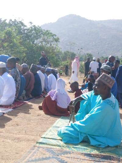 This image depicts an outdoor communal gathering in Kpak, Nigeria, under bright daylight with a mountainous backdrop. Approximately two dozen individuals are visible, predominantly adult males, seated or kneeling on a dry, reddish-brown earthen ground. Many are positioned on small, patterned prayer mats.
In the immediate foreground, right of center, an adult male wearing a light blue embroidered robe and a patterned skullcap sits cross-legged on a green mat, holding a strand of brown prayer beads. His gaze is directed towards the left. Slightly behind him, another individual with their back to the camera wears a light pink top, a reddish-brown skirt, and a white head covering. To their right, a younger person is partially visible.
Further back, multiple rows of men are seated or kneeling, dressed in various traditional attires, including white and dark-colored robes and prayer caps. In the mid-ground, a person in a white *thobe* and a red-and-white checkered headscarf is walking towards the right. Other individuals are standing in the middle distance, some in dark attire.
The environment includes sparse green trees and shrubs. A large, hazy mountain or hill range dominates the background. The ground is dry, open earth.
