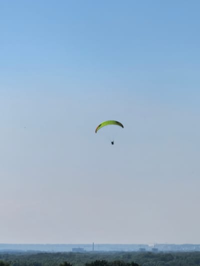 A paraglider is captured in mid-flight against a clear blue sky. The paraglider, a bright green and yellow wing, is suspended above a distant, hazy landscape. The landscape below appears to be a dense forest canopy with faint outlines of industrial structures and construction cranes visible on the horizon, suggestive of an urban or semi-urban area. The scene is calm and serene, with no apparent activity beyond the solitary flight of the paraglider. The location is identified as Naumovka, Russia.