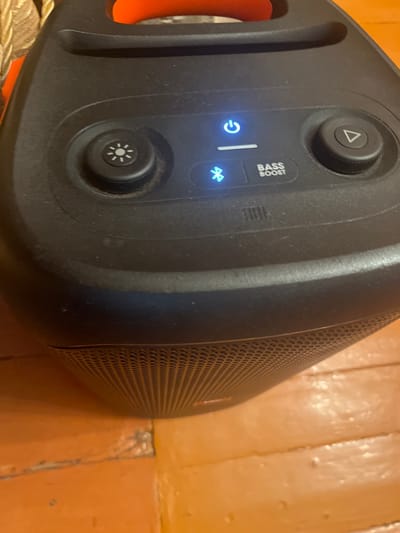The image shows a black portable JBL speaker with an orange handle, placed on an orange wooden floor. The speaker has several illuminated blue buttons: a power button, a Bluetooth symbol, and a "BASS BOOST" label. There are also two rotating dials with symbols on them. One dial has a sun-like symbol, and the other has a play/pause symbol. The speaker's grille is visible at the bottom. The scene appears to be indoors, possibly in a home environment. There are no people or animals in the image. The lighting suggests it might be daytime, but the indoor setting does not provide definitive clues about the specific location within Oktyabrskoye, Russia.