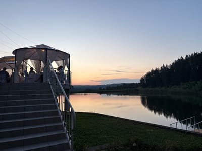 This is a medium-shot, horizontal, eye-level photograph of an outdoor dining area near a body of water at dusk. In the foreground, a concrete staircase with a white metal railing leads up to a large, circular gazebo. The gazebo has a white roof and is draped with white fabric. Inside the gazebo, several people are seated at tables, appearing to be dining. The gazebo is illuminated from within, casting a warm glow. To the right of the gazebo, a wide body of water reflects the soft colors of the twilight sky and the surrounding trees. The water is calm, with only slight ripples. The far bank of the water is lined with dense, dark green trees. The sky transitions from a pale blue at the top to hues of orange and pink near the horizon, indicating sunset. The overall atmosphere is peaceful and serene, suggesting a tranquil evening setting. The location is identified as Skalsko, Bulgaria.