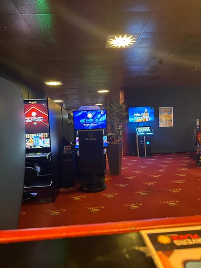 The image depicts the interior of a casino in Beckum, Germany, specifically the "Royal Play" casino. The scene is dominated by rows of brightly lit slot machines, with a red carpet featuring the "Royal Play" logo and crown pattern covering the floor. Several slot machines are visible, including one with a screen displaying "ZONIC" and another with "GIGA GOLDEN NUGGET". To the right, a screen shows "RENTAVIRUS POWER SPINS" next to a poster for "FIVE". The ceiling is dark with a tiled pattern and several star-shaped lights. A large potted plant stands near the center. The overall atmosphere is that of a typical casino environment with flashing lights and entertainment displays.