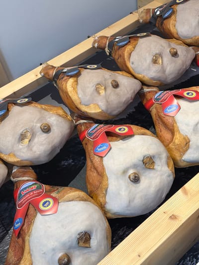 The image displays a close-up, overhead view of several cured hams, likely Prosciutto di Sauris, arranged on a wooden shelf or display in what appears to be a food shop or market. The hams are large, whole, and have a characteristic cured meat appearance with dry, leathery skin and a visible fat cap. Each ham is adorned with red and blue labels that prominently feature the "Wolf" logo and the text "Prosciutto di Sauris" along with other Italian text and seals, indicating the product's origin and quality. Some labels also show a blue oval seal with stars and a circular inscription. The background consists of a grey wall and a dark, patterned surface beneath the hams, possibly a fabric or mat. A light-colored wooden beam runs diagonally across the lower right portion of the frame, suggesting part of the shelving unit. The lighting is even and illuminates the details of the hams and their labels. The scene is static, with no people or actions depicted. The overall impression is one of a well-presented food product display in Sauris, Italy, a town renowned for its unique cured ham.