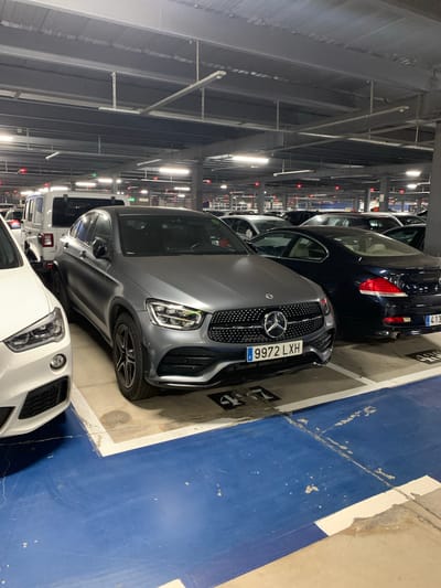 In the city of Manises, Spain, inside a multi-story parking garage, a grey Mercedes-Benz GLC Coupe with license plate "9972 LXH" is parked between a white vehicle and a dark blue coupe. The Mercedes features a distinctive grille with the Mercedes-Benz logo. A white Jeep is parked behind the Mercedes. The parking garage has a concrete ceiling with exposed lighting fixtures and a red indicator light above each parking spot. The floor is painted blue and beige with white dividing lines.