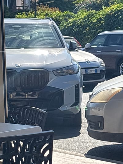 A light-colored BMW SUV is parked on a street in Ardea, Italy. The car is in the foreground, with its front grille and headlights visible. A dark-colored SUV is parked behind the BMW, and a lighter-colored sedan is parked to the right. The street is paved, and there are trees and bushes in the background. The license plate of the BMW is visible and reads "EX-473TE".