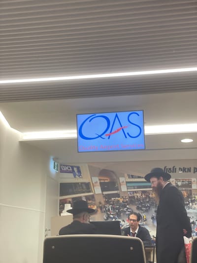 The image depicts an indoor scene within an airport in Israel, specifically at a "Quality Airport Services" counter. The setting features a modern ceiling with linear lighting and acoustic panels. A large digital display screen, centrally mounted, shows the blue "QAS" logo with a red underline and the red text "Quality Airport Services." Below and behind the screen, a large mural or wallpaper depicts a busy outdoor scene, possibly a marketplace, with Hebrew text elements. To the left, a green exit sign with Hebrew characters is visible on a white wall.
In the foreground and midground, three adult males are present. On the left, a man dressed in a black fedora and dark suit, consistent with Haredi Jewish attire, is seated at a desk, largely seen from the back, appearing to engage with a computer monitor. Next to him, a man wearing glasses, a dark jacket, a white shirt, and a red tie is seated at the same desk, looking towards the right. On the right, a third man, also in a black fedora and dark suit with a prominent reddish-brown beard and peyos, stands facing left towards the desk. He holds a small, colorful bag in his right hand, with his left hand behind his back. The interaction suggests a service or transaction occurring at the desk.
