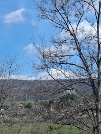 The image depicts a scene in Gradets, Bulgaria, featuring a mountain range with a visible cliff face running along the middle of the frame. The lower slopes of the mountain are partially forested, with occasional buildings and power lines visible. In the foreground, bare tree branches frame the mountain and a blue sky with scattered white clouds. The perspective is slightly angled, capturing both the sky and the terrain below.