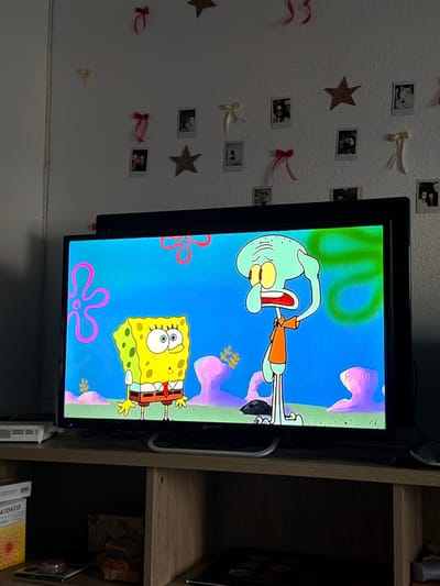 A television set displays a scene from the animated series SpongeBob SquarePants, featuring SpongeBob and Squidward. The TV is positioned on a wooden media console. The background of the scene shows a vibrant blue underwater environment with pink sea anemones. The wall above the media console is decorated with various small framed photographs, glittery stars, and decorative bows. The overall setting appears to be a domestic interior, possibly a living room or bedroom, in Castillo Bajo-conejito, Spain.