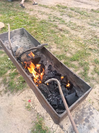 This media file shows a close-up view of a charcoal grill with a fire burning in it. The grill is a long, rectangular metal container filled with charcoal and burning logs. Flames are visible, indicating that the fire is active. A metal poker rests inside the grill, suggesting someone is preparing to cook.
In the background, the scene transitions to an outdoor setting with dry, cracked earth and sparse green grass. Two people's legs are visible walking away from the grill, wearing casual footwear. One person is wearing dark sandals with straps, and the other is wearing flat, black shoes. The presence of the grill, the outdoor environment, and the people walking suggests a casual outdoor gathering, possibly a barbecue or picnic.
The time of day appears to be daytime, as indicated by the natural lighting. The weather seems to be fair, with no visible rain or strong winds. The ground condition suggests a dry climate or season. The location cues of dry earth and grass are consistent with a rural or semi-rural setting, potentially in Toguchin, Russia, as suggested by the context. No text is visible in the image.