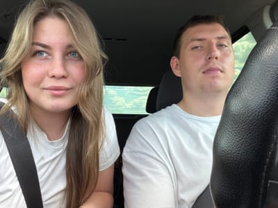 Two people, a woman and a man, are seated in the front seats of a vehicle. The woman, on the left, has long blonde hair and blue eyes, with light skin and freckles. She is wearing a white t-shirt and a black seatbelt. The man, on the right, has short brown hair, blue eyes, and fair skin. He is wearing a white t-shirt. The interior of the vehicle is visible, including a black steering wheel and dark headliner. Through the window behind them, a cloudy sky is visible. The scene suggests a daytime setting, likely during a car journey. No specific location within Alnashi, Russia is identifiable from the image.