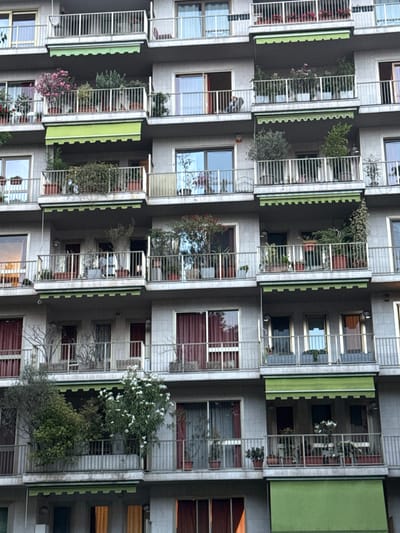 The image is an exterior, medium shot of an apartment building in Sesto San Giovanni, Italy. The building is several stories high, with multiple balconies on each level. The balconies are lined with metal railings and have potted plants and trees. Some balconies have awnings. The building has a light-colored facade. There are no people clearly visible in the image. The lighting suggests it is daytime, possibly overcast.
