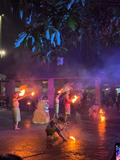 The image captures a nighttime fire performance occurring outdoors, likely within the Sunway Lost World of Tambun theme park in Ipoh, Malaysia. The scene is illuminated by the intense orange glow of multiple fires and strong blue-purple stage lighting, which casts vibrant reflections on the dark, reflective ground.
Four individuals, identified as performers, are actively engaged with fire. On the left, a barefoot female performer in a light-colored top and patterned skirt holds two flaming staffs horizontally. To her right, another performer, wearing a red top, patterned skirt, and a headpiece, stands with multiple flaming objects. In the foreground, a performer kneels, manipulating a flaming prop on the ground. Further right, another performer also kneels near ground-level flames. Their attire consists of light, tropical-themed skirts or wraps.
A large, textured, brown statue resembling a tiki figure with prominent facial features in red and white stands centrally on the performance area. Several smaller, tripod-mounted flames are positioned behind the performers. Overhead, large-leafed trees with hanging vines are visibly illuminated by the blue stage lights. Haze or smoke from the fires permeates the air.
In the background, behind structural pillars and overhead beams, an audience is visible, composed of multiple individuals seated and standing, observing the performance. The bottom right corner shows a partial view of a smartphone screen, indicating the image was captured by an audience member.