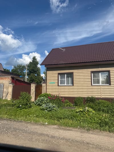 A residential building with beige siding and a dark brown roof is situated in the foreground, to the right of the frame. Two white-framed windows with white curtains are visible on the building's facade. A street sign with Cyrillic text, "ФАБРИЧНАЯ 14," is affixed to the wall. A lush garden with various green plants and some pink flowers borders the house. To the left of the house, there is a brick fence with a dark brown metal gate. Behind the fence, a smaller red building with a metal roof is visible, along with some trees and utility poles. The scene is set under a bright blue sky with scattered white clouds. The ground in front of the property is a dirt road. The image appears to be taken in Medyn, Russia.