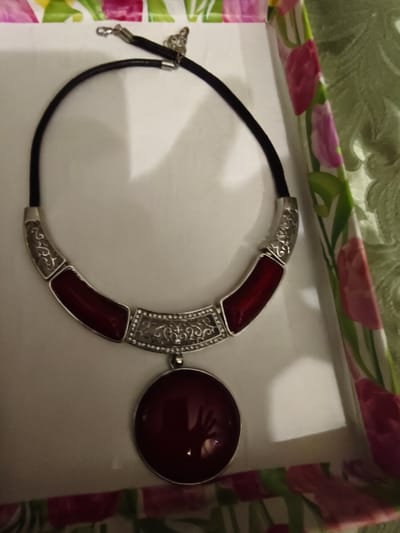 The image shows a close-up, overhead view of a necklace lying on a white surface, possibly inside a box. The necklace has a black cord and features a decorative silver-toned centerpiece. This centerpiece is composed of curved sections with intricate cut-out designs and inlays of a deep red, semi-precious stone. A large, circular, deep red pendant hangs from the center of the necklace. The necklace is resting on a white, slightly creased surface, and the edges of a decorative box with a floral pattern of pink and green tulips are visible on the sides and bottom of the image. The lighting suggests an indoor setting with a soft, diffused light source. There are no people or animals in the image, and no specific location within Bolshaya Martynovka, Russia, can be identified.
