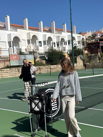 The image depicts a bright, sunny outdoor scene on a green tennis court in Ericeira, Portugal. In the foreground, positioned to the right of a black tennis net with a white top band, stands a female with long, wavy brown hair. She wears a grey knit hooded sweater, light beige wide-leg ribbed pants, and light-colored sneakers. Her face is partially obscured by her hair, and sunglasses hang from the front of her sweater. Adjacent to the net is a black wheeled ball cart containing yellow tennis balls, a black cap with "arc" text, and a black bag with a white circular logo featuring intersecting lines. A larger black banner with white vertical lettering and a circular logo is positioned behind the cart.
In the midground, to the left of the net, a blonde female wearing a black top and plaid pants stands on a dark, flat board, possibly a skateboard. She is facing towards the center-right. Behind her, a male wearing a light-colored baseball cap, grey hoodie, and dark pants holds a brown, wooden-appearing tennis paddle or training racket. A yellow tennis ball lies on the court surface near the blonde female.
In the far background, beyond a green wire fence and a stacked stone wall, a male figure is visible, engaged in playing tennis on another part of the court or an adjacent court. He wears a dark shirt and shorts and holds a tennis racket.
The background is dominated by a row of white residential buildings with terracotta tiled roofs, multiple arched balconies, and distinctive tall, curved chimneys, characteristic of Portuguese architecture. Vegetation, including bushes and small trees, is visible along the stone wall and in front of the buildings. A banner with "tennis school" and other logos is visible on the fence. The sky is clear and blue, and the scene is illuminated by strong sunlight, casting visible shadows.