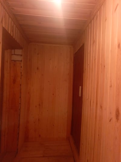 The image depicts an interior view of a wooden sauna. The walls and ceiling are paneled with light-colored wood, and the floor is also wooden. A dark-colored door with a light-colored handle is visible on the right side of the room. On the left side, a section of exposed brick wall is visible behind a wooden panel. The lighting appears to be artificial and casts a warm glow throughout the space. There are no people or animals visible in the image. The scene is consistent with a sauna located in Bogatye Saby, Russia.