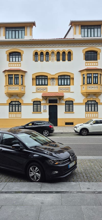 This image captures a street scene in Póvoa de Varzim, Portugal, featuring a white building with yellow architectural accents and several parked cars. The building, characterized by its ornate windows, bay windows, and decorative frieze, is a prominent element. In the foreground, a black Volkswagen Polo is parked at an angle on a sidewalk paved with large grey tiles and cobblestones. Behind it, a black SUV is parked further down the street, and a white Nissan Juke is parked on the right side of the image. The street is paved with asphalt. The sky is overcast.