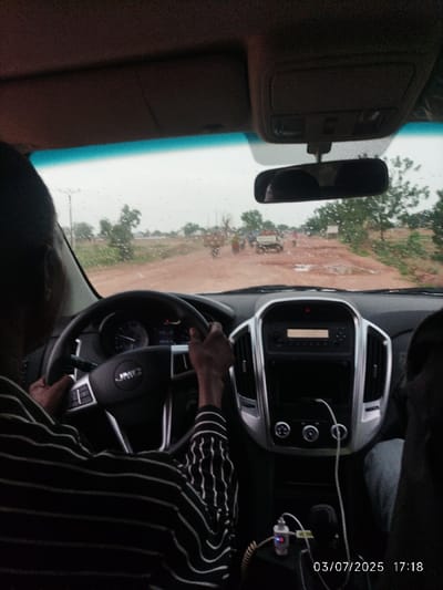 Inside a vehicle, a person with dark skin drives while gripping a steering wheel with a "JMC" logo. The dashboard has a radio unit and climate controls. Outside, the view through the windshield shows a dirt road with puddles, buildings, trees, and people. Raindrops are visible on the glass. A rearview mirror is positioned above the dashboard. The bottom right corner displays a date and time stamp. The driver is wearing a striped shirt.